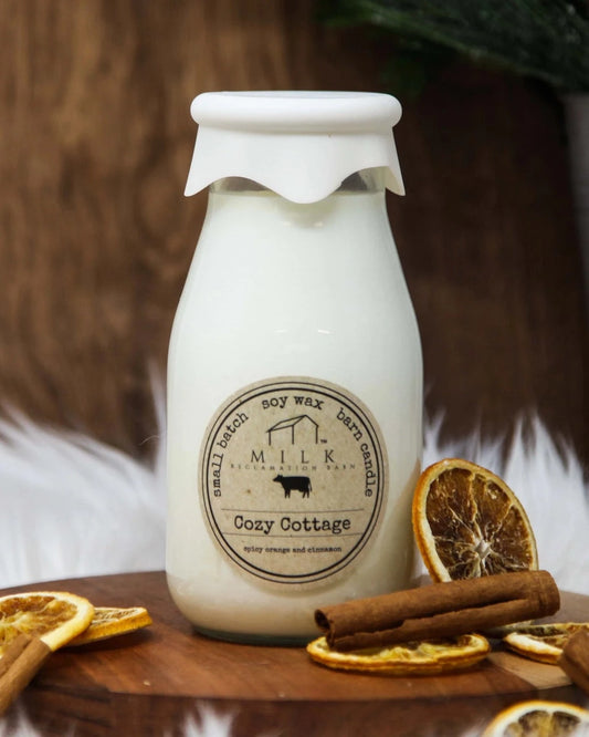 Milk bottle-shaped candle labeled 'Cozy Cottage' on a wooden surface with dried oranges and cinnamon sticks.