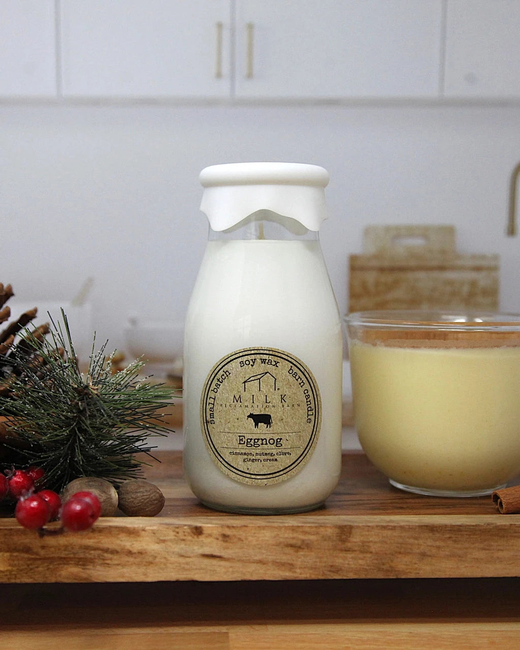 Milk bottle labeled 'Eggnog' next to a glass of eggnog on a wooden surface with Christmas decorations.
